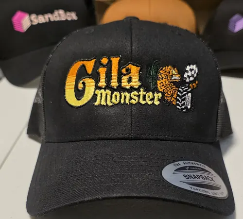 Close-up of a black Yupoong trucker with the gold Gila Monster logo and lizard